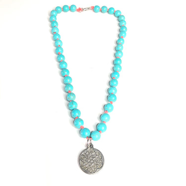 Collar corto de piedra natural Howlite en color turquesa, las cuentas están unidas en hilo fucsia. Medallón y cierre en zamak bañado en plata.